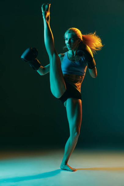 Jedna młoda kobieta, zawodowa kickboxer w mundurze sportowym i trening rękawiczek odizolowana na ciemnozielonym tle w neonie. Sport, konkurencja, koncepcja władzy. - Zdjęcie, obraz