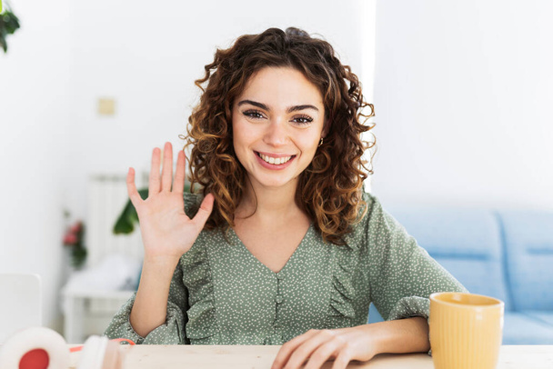 Pretty young Hispanic, vlogger woman with curly hair waving hand looking at webcam talking to camera sits at table, video call, recording blog, headshot portrait. - Foto, afbeelding