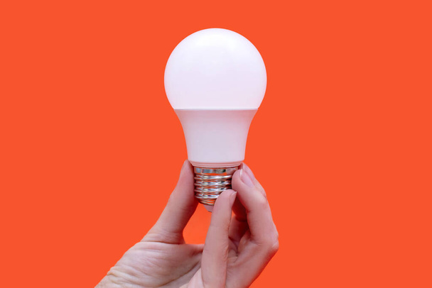 Energiesparlampe in der Hand auf orangefarbenem Hintergrund. Energiesparen zu Hause. Elektrische Geräte und die Entwicklung des Lichts. Naturschutz und Umweltschutz - Foto, Bild