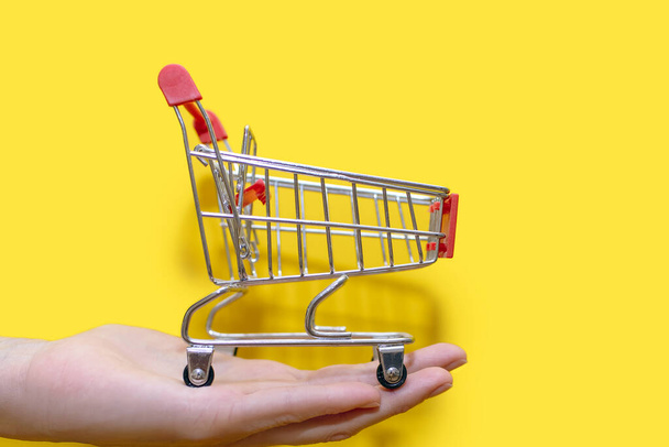Small shopping trolley in hand on a yellow background close-up. The concept of buying and selling. Delivery of goods to the trading floors. A small toy cart with wheels. Empty shopping cart - Zdjęcie, obraz