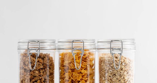 food storage, healthy eating and diet concept - close up of jars with oat, corn flakes and granola on white background - Φωτογραφία, εικόνα