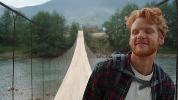 Positive traveler hiking mountains outdoors on river bridge. Portrait of happy redhead travel nature landscape. Curious man look around forest. Young backpacker walk on summer vacation. Active concept - Metraje, vídeo