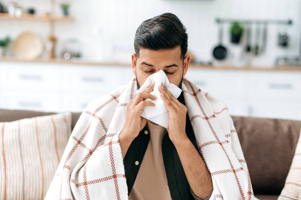 Nezdravý indický nebo arabský chlap sedí doma na pohovce v obývacím pokoji pod kostkovaným, drží ubrousek v blízkosti nosu, kýchá, trpí rýmou, necítí se dobře, potřebuje léčbu a konzultace s lékařem - Fotografie, Obrázek