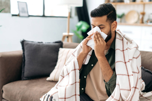 Sneezing, runny nose, cough. Ill arabian or indian young man, sits on the couch at home under a plaid, sneezing in a napkin, feels weak, unwell, needs treatment and medicines - Photo, image