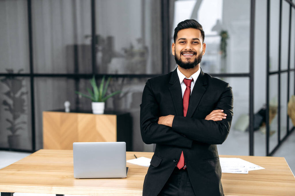 Pozytywnie przystojny, pewny siebie, odnoszący sukcesy indyjski lub arabski biznesmen, w garniturze, męski przedsiębiorca, stoi w nowoczesnym biurze przy biurku z skrzyżowanymi ramionami, patrzy w kamerę, uśmiecha się przyjazny - Zdjęcie, obraz