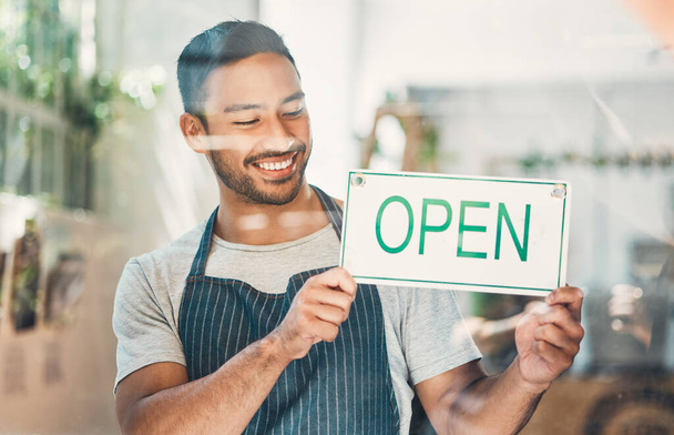 One young hispanic man hanging up an open sign in at a window on display in a cafe or store. Happy mixed race guy excited to open his shop and welcome customers. - Foto, immagini