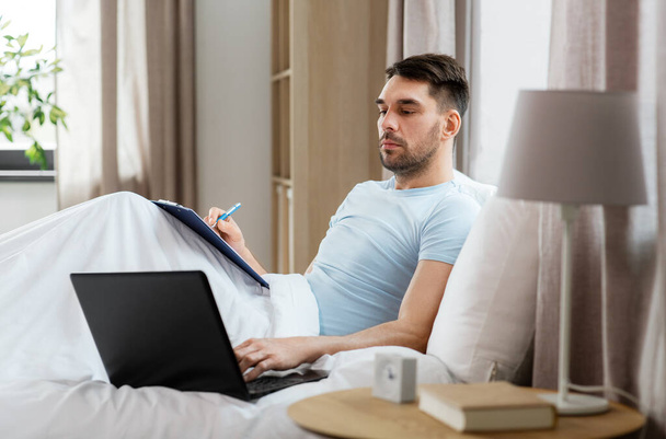 people, technology and remote job concept - man with laptop computer and clipboard working in bed at home bedroom - Фото, зображення