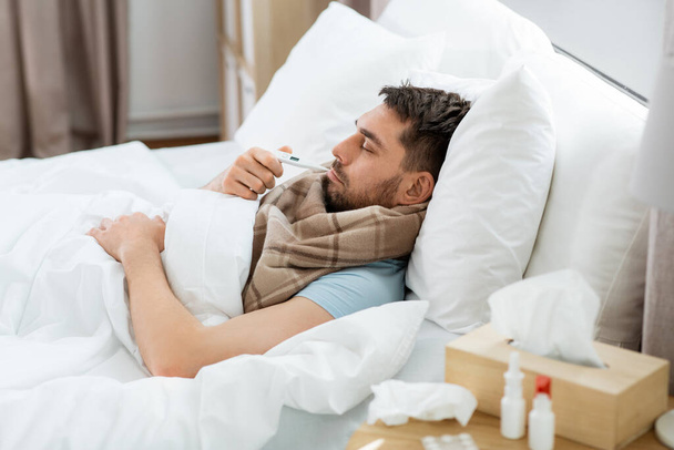 people, health and fever concept - sick man in scarf measuring temperature by thermometer lying in bed at home - Foto, Bild