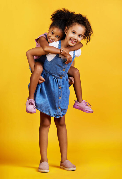 Mixed race girl piggybacking her sister, sibling or friend in studio isolated against a yellow background. Cute hispanic children posing inside. Happy and carefree kids playing together and bonding. - Foto, imagen