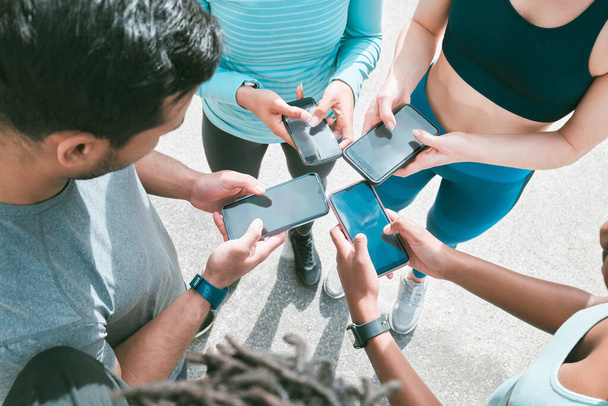 Closeup of diverse group of sporty people from above using cellphones with blank screens in synchronicity while standing in a circle around each other. Hands of athletes browsing online and searching - Foto, immagini