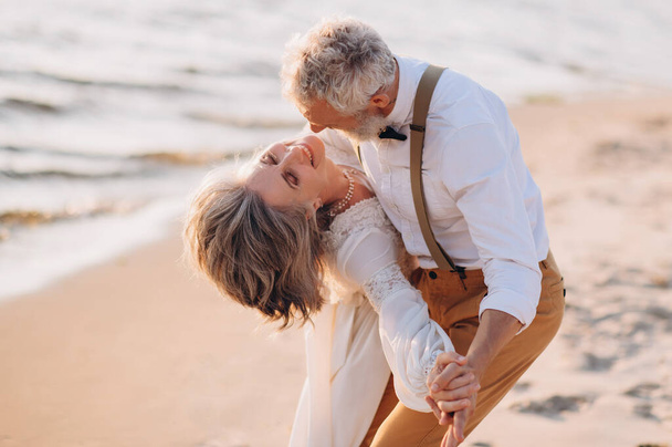 Happy elderly couple dancing on the beach on a sunny day. The groom dances with the bride on the wedding day. - Foto, imagen