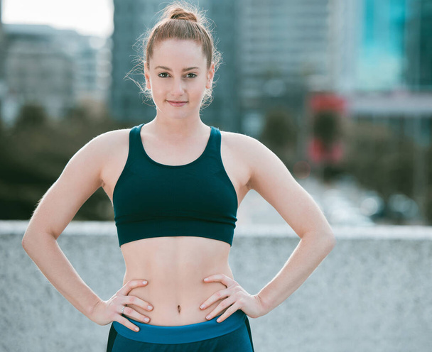 Portrait of one confident young caucasian woman standing with hands on hips ready for exercise outdoors. Determined female athlete looking focused and motivated for training workout in the city. - Foto, Imagen