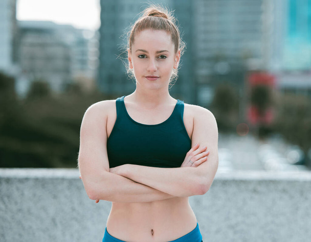 Portrait of one serious young caucasian woman standing with arms crossed ready for exercise outdoors. Determined female athlete looking focused and motivated for training workout in the city. - 写真・画像