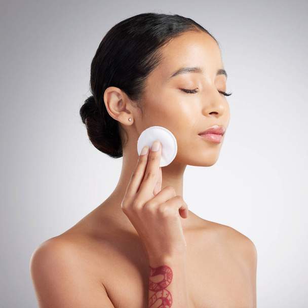 A beautiful mixed race woman using a cotton pad to remove makeup during a selfcare grooming routine. Hispanic woman applying toner to her face against grey copyspace background in a studio. - Fotoğraf, Görsel