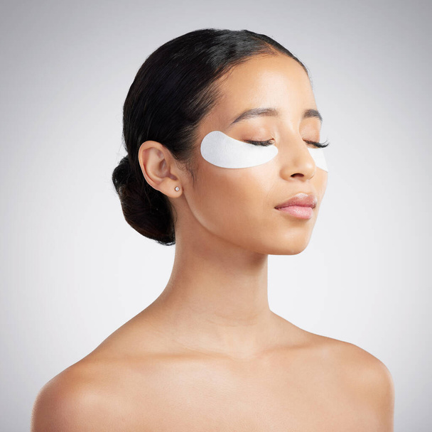A beautiful mixed race woman wearing under eye patches. Hispanic model with glowing skin using hydrating treatment against a grey copyspace background. - 写真・画像