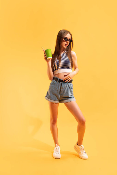 Tasting lemonade. Studio shot of emotive young girl isolated on yellow color background. Concept of beauty, art, fashion, emotions, youth, summer fashion, vacation. Copy space for ad - Фото, зображення