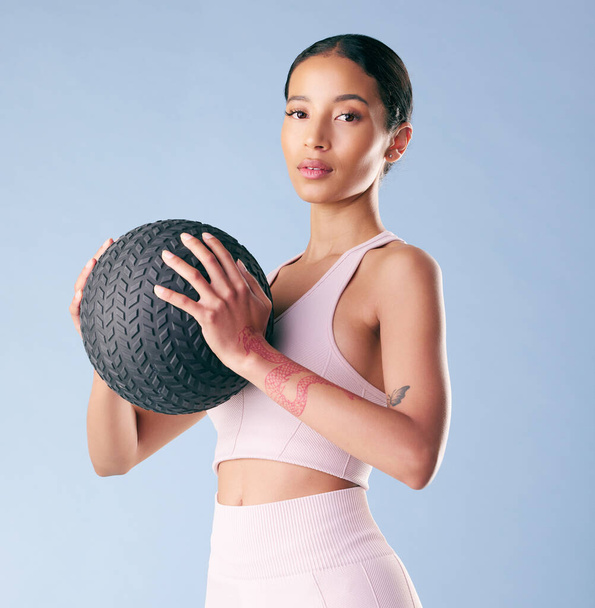 Mixed race fitness woman standing with a medicine ball or slam ball in studio against a blue background. Beautiful young hispanic female athlete exercising or working out. Health and fitness. - 写真・画像