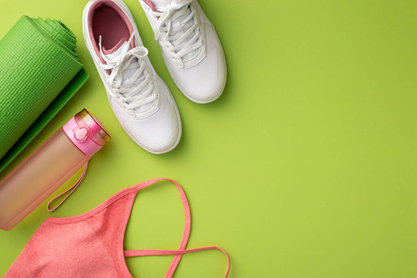 Fitness concept. Top view photo of white sneakers pink sports top bottle of water and green exercise mat on isolated green background - Foto, imagen