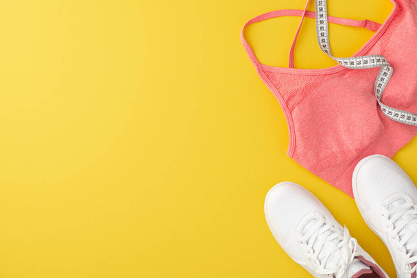 Спортивная концепция. Фотография сверху розового спортивного бюстгальтера, белые туфли и лента на изолированном желтом фоне с копирайтом - Фото, изображение