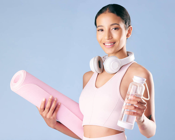 Mixed race fitness woman standing with her yoga mat and water bottle in studio against a blue background. Beautiful young hispanic female athlete exercising or working out. Health and fitness. - Foto, Imagem