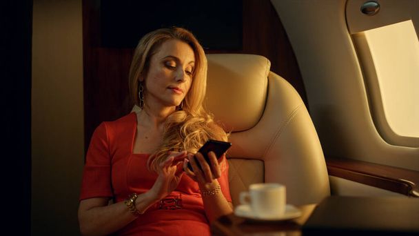 Usmívající se pasažér mluví smartphonem ve zlatém slunečním světle. Nádherná žena odpočívající mluvit mobilní telefon s rodinným partnerem na luxusní výlet. Elegantní obchodnice cestuje soukromým letadlem. Koncept bohatého životního stylu - Fotografie, Obrázek