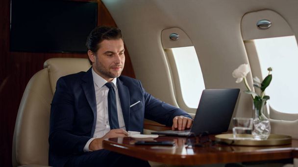 Soustředěný obchodník, který vypadá jako počítač na služební cestě. Bohatí muži pracují na notebooku a kontrolují online obchodní zprávy. Úspěšný manažer vytvoří prezentaci v okně letadla. Mužský profesionální koncept. - Fotografie, Obrázek