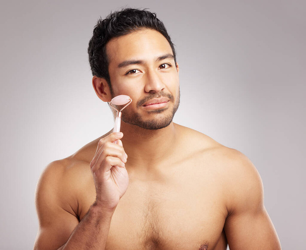 Handsome young mixed race man posing shirtless in studio isolated against a grey background. Hispanic male using a dermal face roller to massage and relax his skin. All part of his skincare regime. - Foto, imagen