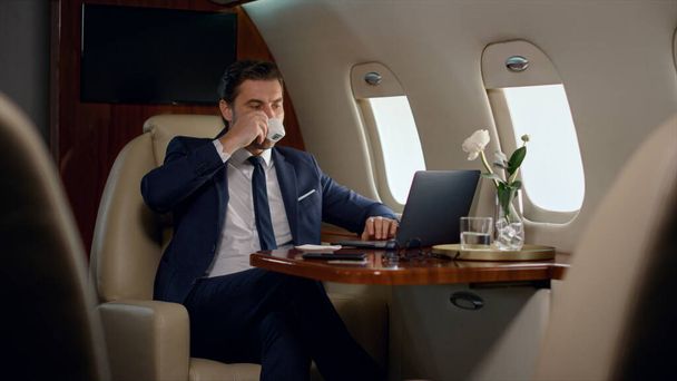 Finance manager take sip of coffee on business trip. Confident boss work laptop checking marketing sales statistics on luxury private jet. Focused stylish man looking airplane window drinking beverage - Photo, Image