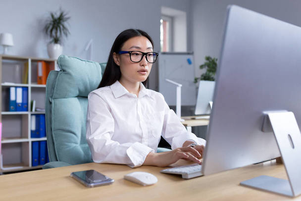Asijská podnikatelka pracující v moderní kanceláři na počítači, psaní online sestavy. - Fotografie, Obrázek