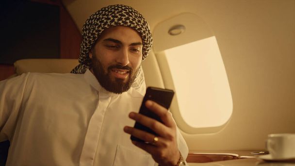 Successful man surfing web at airplane window closeup. Arabian hold smartphone enjoying social media content in private jet. Cheerful bearded guy read news checking email. Business travel concept - Photo, Image
