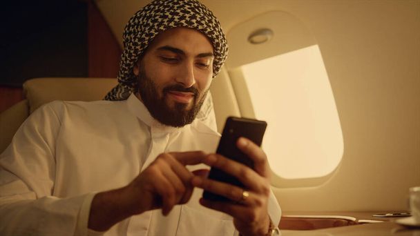 スマートフォンのクローズアップでアラビア語を笑顔。ビジネス旅行で手に電話をスワイプする陽気な男。豊かな成功した会社の所有者は、伝統的な衣装で電子メールのソーシャルメディアをチェックします。ソーシャルネットワーキングコンセプト. - 写真・画像