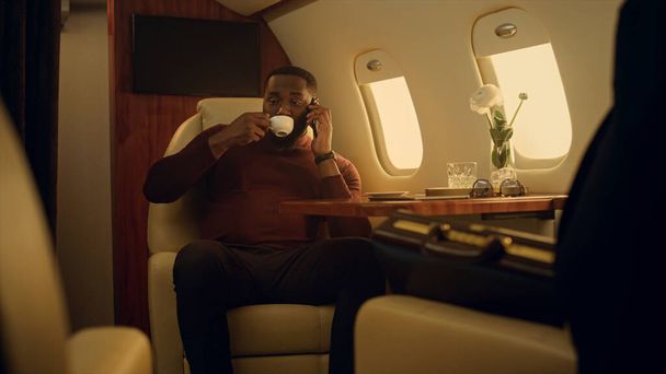 アフリカ系アメリカ人が出張中にスマートフォンを話す。一等航空機で休んで旅行を楽しんで楽しい男。パートナーとのコミュニケーションに成功した黒髪のビジネスマンコーヒーを飲む. - 写真・画像