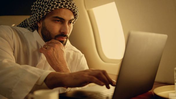 Bedachtzame moslim werkende laptop op zakenreis close-up. Rijke man typt bedrijfsstatistieken bij vliegtuigraam. Pensief zelfverzekerde zakenman overweegt project in traditionele kleding. - Foto, afbeelding
