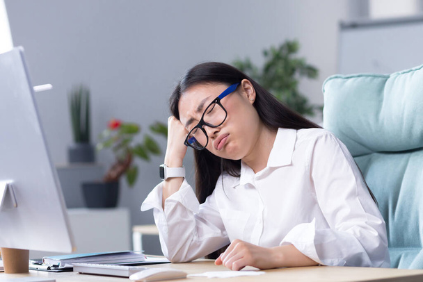 A sad Asian business woman despairs of her abilities, is depressed and works in the office at the computer. - Foto, imagen