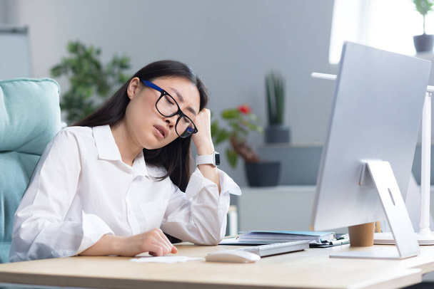 A sad Asian business woman despairs of her abilities, is depressed and works in the office at the computer. - Фото, изображение