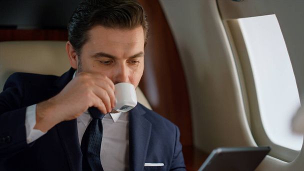 Knappe zakenman met een tablet aan het raam. Een close-up man die op zakenreis is. Gericht stijlvolle manager drinken koffie aanraken digitale pad scherm op vliegtuig. Succesvol ondernemersconcept. - Foto, afbeelding