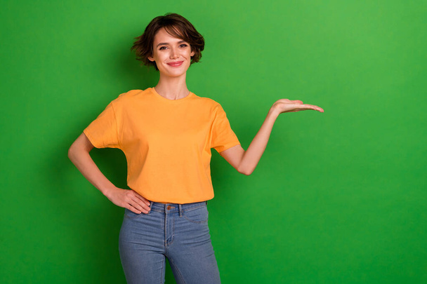 Photo of sweet funny young lady wear orange outfit smiling holding arm empty space isolated green color background. - Zdjęcie, obraz