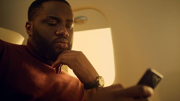 Focused guy looking smartphone at airplane window closeup. Hands holding cell phone. Serious ceo work checking stock market data. Pensive nervous african american browse web. Male professional concept - Photo, Image