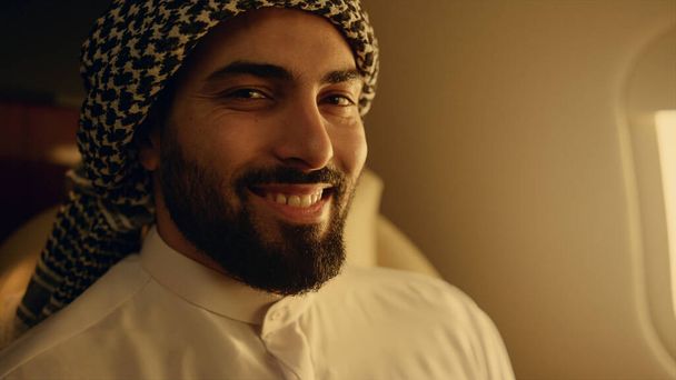 Smiling arabic man posing at airplane window closeup. Happy businessman travel luxury first class in traditional outfit. Attractive bearded company owner resting enjoying business trip. Wealthy people - Photo, image