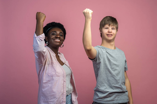 Team. African american woman and caucasian guy with down syndrome with raised fist looking at camera standing on light background - Φωτογραφία, εικόνα