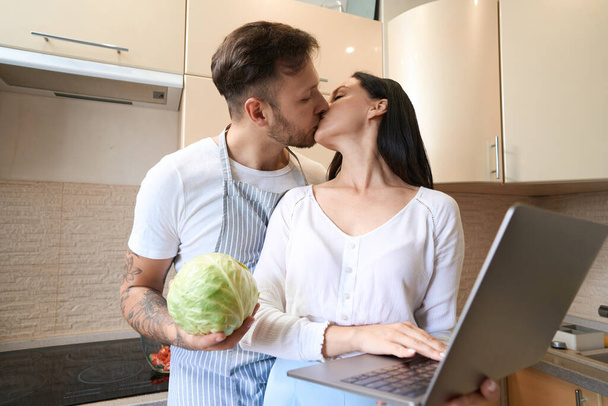 Man with cabbage giving his romantic partner a kiss on mouth while she clicking laptop keyboard in kitchen - Fotó, kép