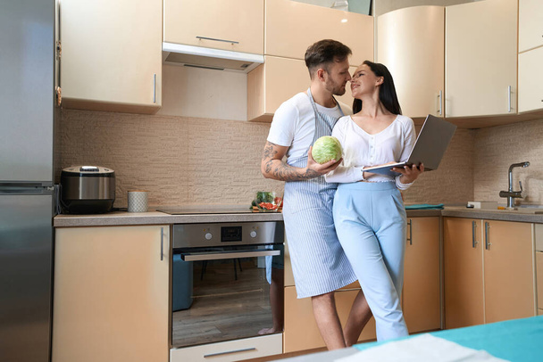 Joyful lady smiling at her boyfriend with cabbage in kitchen while standing beside him with laptop - Φωτογραφία, εικόνα