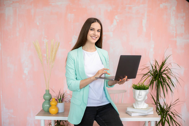 Retrato de menina elegante alegre em roupa casual inteligente em pé na mesa segurando laptop nas mãos. Foto de alta qualidade - Foto, Imagem