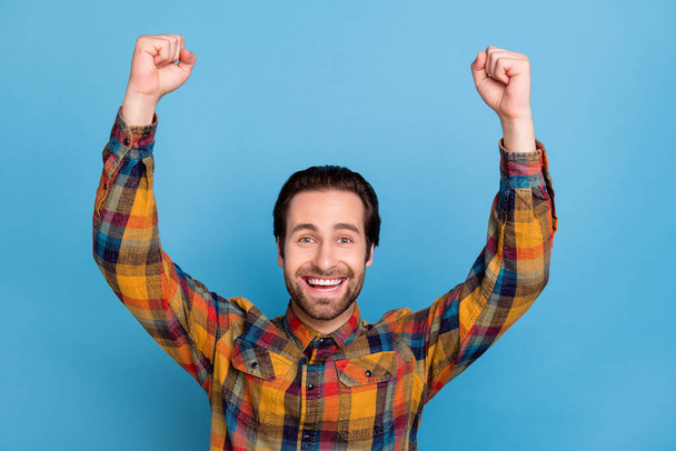 Photo of positive lucky young man wear plaid shirt smiling rising fists isolated blue color background. - Foto, imagen