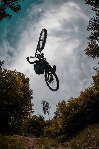 Σιλουέτα ενός ποδηλάτη βουνών πηδώντας πάνω από την κάμερα και εκτελώντας μαστίγιο ουρά. Ακραία φωτογραφία του mtb racer άλμα που περιβάλλεται από δέντρα και συννεφιασμένο ουρανό. Καλό ιστορικό και πλάνο δράσης. - Φωτογραφία, εικόνα