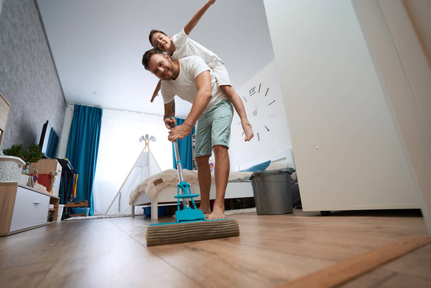 Parent using microfiber mop for cleaning floor in bedroom with child spreading arms while sitting on his back - Foto, imagen