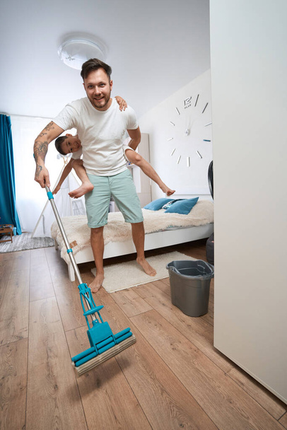 Excited father carrying boy on his back in bedroom while using mop for cleaning floor with water - Photo, image