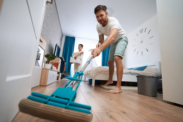 Male parent washing room floor with microfiber mop while child gathering baskets and boxes in background - Foto, immagini