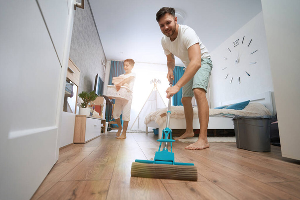 Kid bringing boxes in his hands through bedroom while man using mop for washin floor from dirt - Zdjęcie, obraz