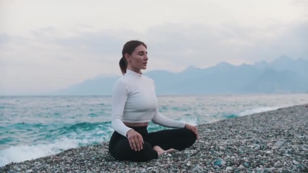 Eine Frau in weißem Pullover sitzt in Lotusposition am Kiesstrand am Meer. Mittelschuss - Filmmaterial, Video